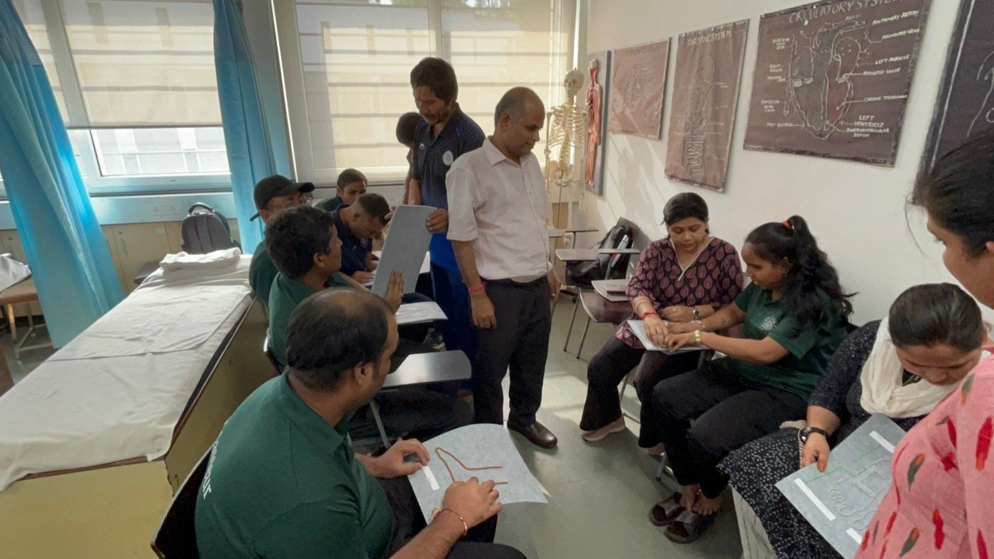 The acupressure class at the Blind Relief Association mid-session: an instructor stands at the centre holding a tactile placard while learners in green BRA polo shirts work through raised-relief diagrams on their laps, anatomy charts and a skeleton behind them.