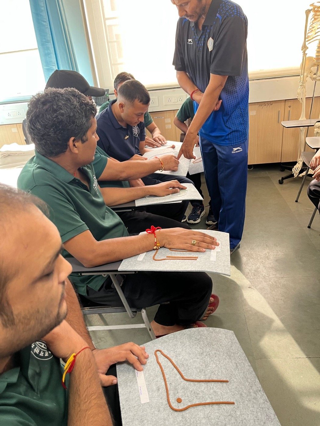 An instructor at the Blind Relief Association leaning in to point out a feature on a learner&rsquo;s tactile diagram, while others in the row read their own raised-relief sheets.
